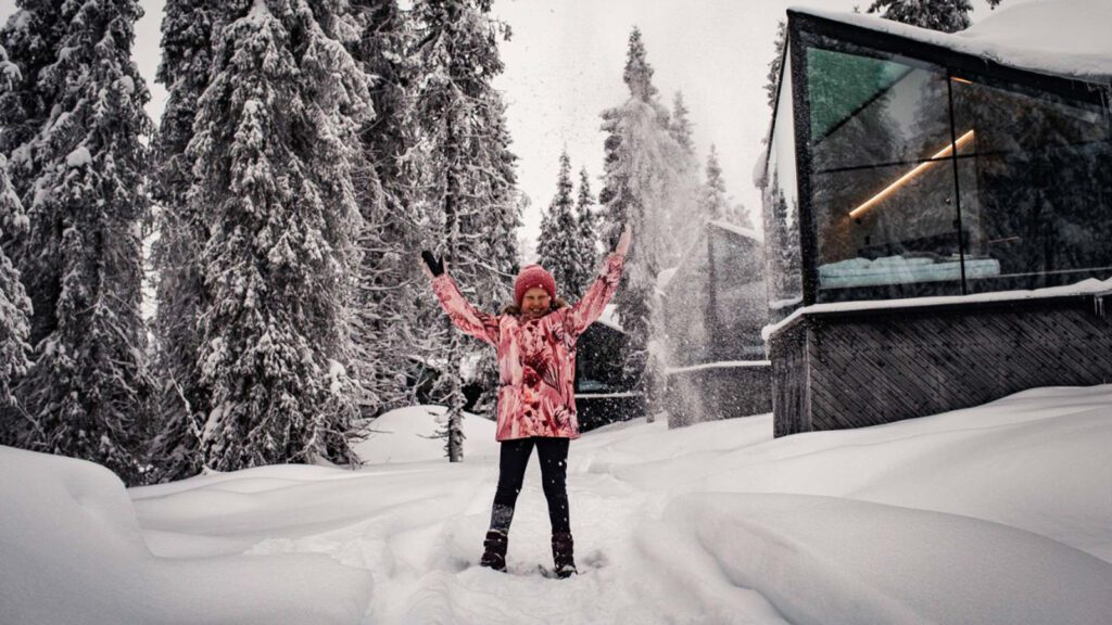 Young girl in a coat playing in the snow with trees and huts in the background